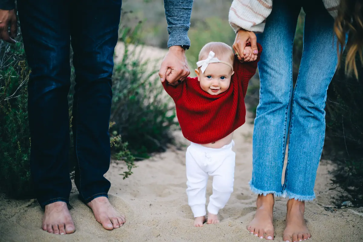 Baby in a red sweater walking barefoot in the sand, holding her parents’ hands