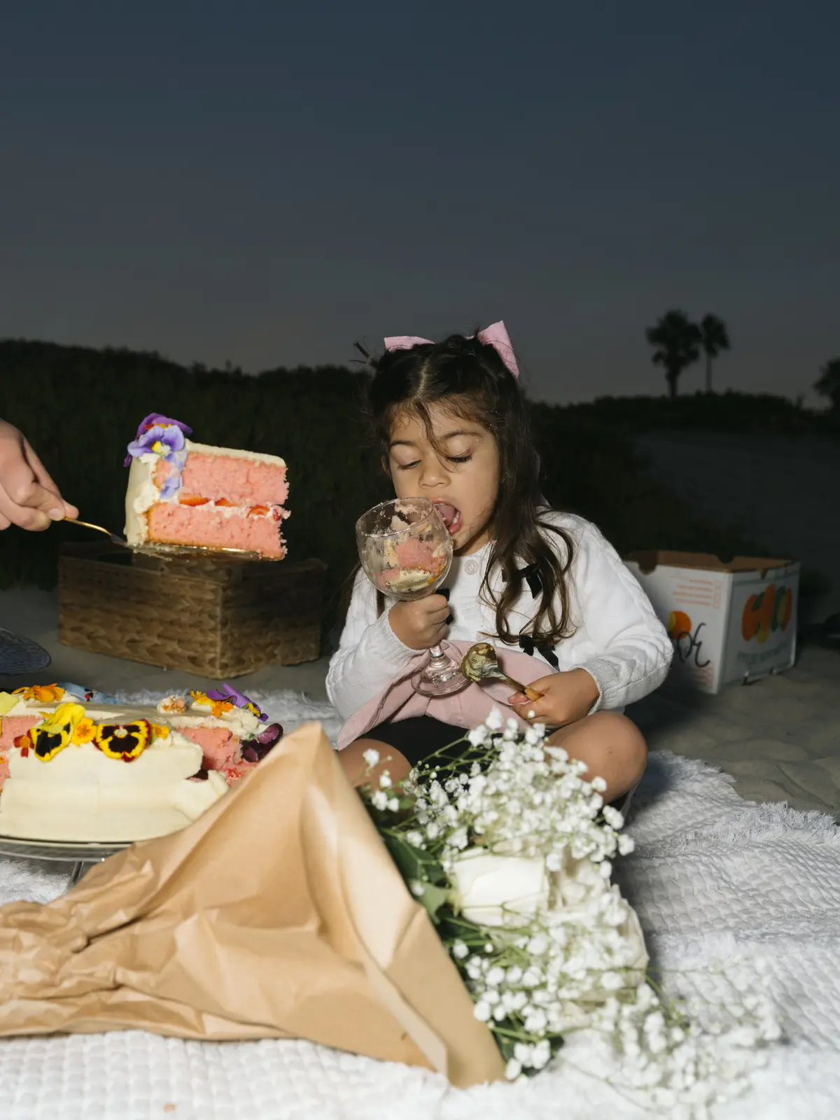 Little girl in a pink bow licking frosting from a glass at a twilight beach birthday picnic with cake and flowers