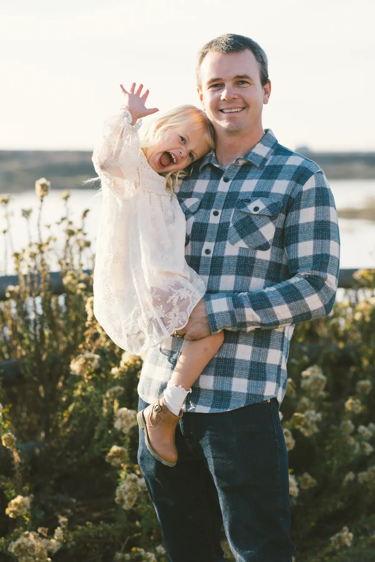 Dad holding his daughter as she laughs and waves, with the bay behind them