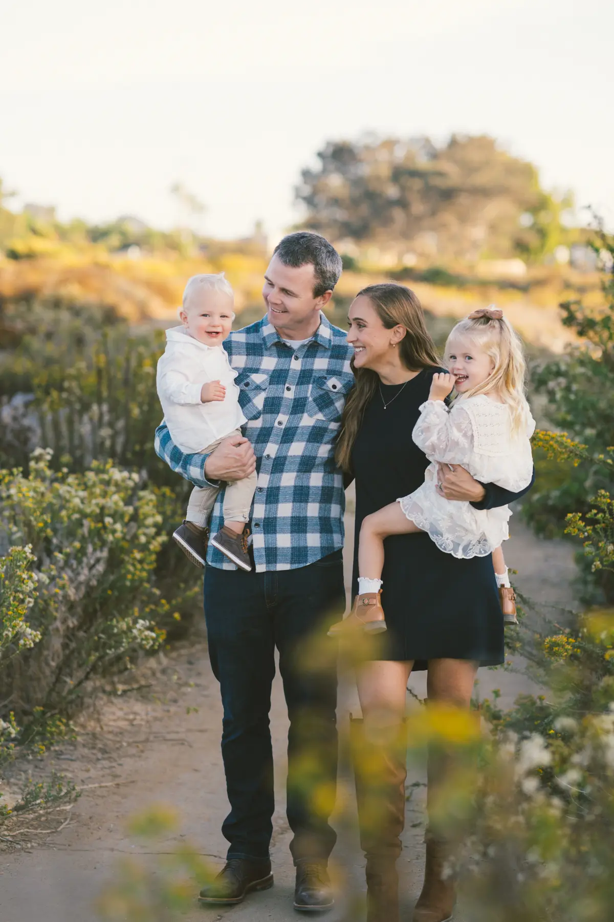 Family of four smiling together among yellow wildflowers on a coastal trail