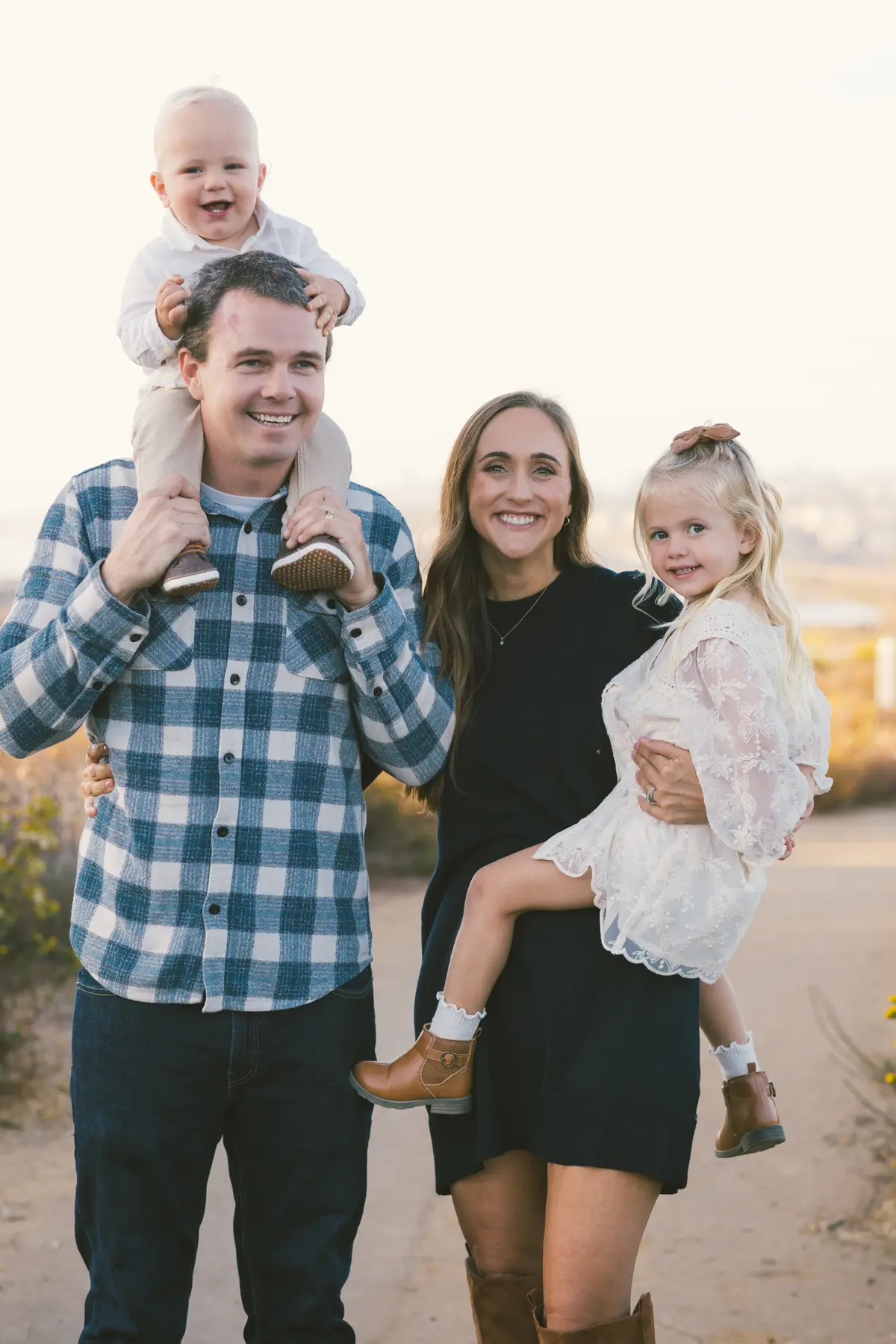 Baby riding on dad’s shoulders beside mom and big sister at golden hour