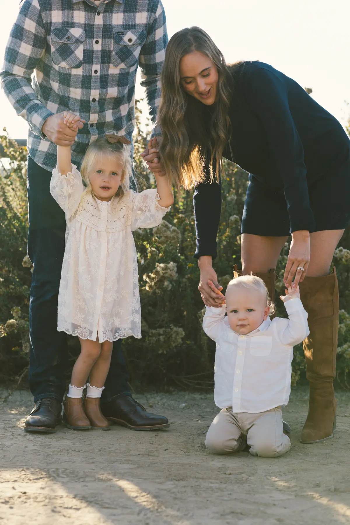 Big sister and baby brother holding their parents’ hands on a sandy trail