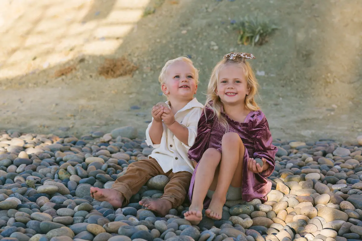 Brother and sister sitting barefoot on beach cobblestones in their holiday outfits